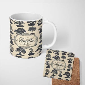 May include: A white ceramic mug and a square cork coaster, both featuring a cream-coloured background with a repeating pattern of dark blue bonsai tree silhouettes and a central decorative frame containing the name Amelia in script font.