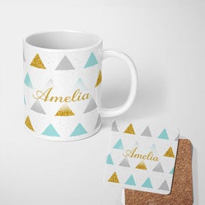 May include: A white ceramic mug and a square coaster featuring a repeating pattern of gold glitter, grey, and light blue triangles on a speckled white background. The name Amelia is printed in gold cursive script on both the mug and the coaster.