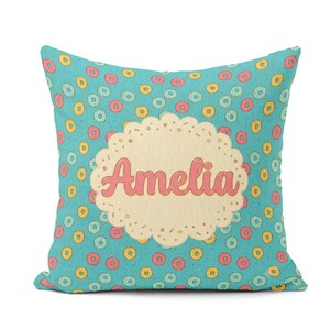 May include: A square throw pillow with a teal background featuring a repeating pattern of small pink, yellow, and light blue doughnuts. In the centre, a cream-coloured scalloped circle contains the name Amelia written in a pink script font.