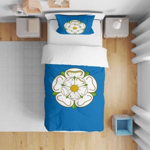 May include: A blue duvet cover and pillowcase set with a white rose emblem, yellow center, and green details. The bedding is on a bed with a white top sheet. The set is designed for a single bed.