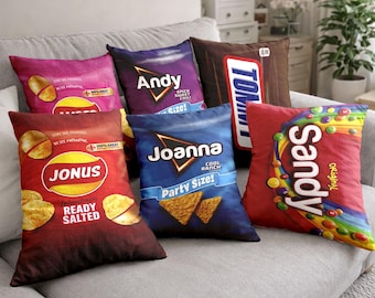 Op de afbeelding: Een collectie van vijf vierkante sierkussens met grafische prints van snackverpakkingen. De ontwerpen omvatten Jonus Ready Salted, Andy Spicy Sweet Chili, een bruine chocoladereepverpakking, Joanna Cool Ranch en Sandy Original met kleurrijke snoepjes.