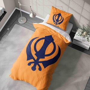 May include: Bedding set in orange and navy blue, featuring the Khanda symbol. The set includes a duvet cover and a pillowcase. The bedding is displayed on a bed with a white sheet. The room has neutral tones.