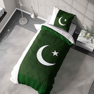 May include: A green and white bedding set featuring the flag of Pakistan. The duvet cover and pillowcase display a crescent moon and star symbol. The bedding is on a white bed, with a gray rug and a lamp in the background.
