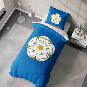 May include: Blue duvet cover and pillow set with a white rose emblem, yellow centre, and green leaves. The bedding is on a white mattress. The duvet cover measures approximately 135cm x 200cm.