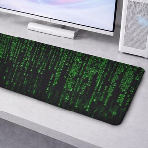 May include: A black rectangular desk mat with a green digital rain design. The mat is on a light grey surface, with a computer monitor and tower in the background. The pattern resembles computer code, a tech-themed accessory.