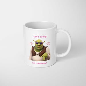 May include: White ceramic mug with a cartoon image of Shrek wearing sunglasses and a brown vest. The mug has the text "can't today, I'm swamped" in pink, with pink heart accents. The mug has a white handle.