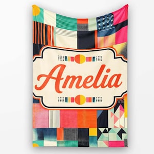 May include: A rectangular throw blanket featuring a vibrant patchwork of geometric patterns in shades of red, orange, yellow, teal, blue, and black. A central cream-coloured label with a dark border displays the name Amelia in red script.