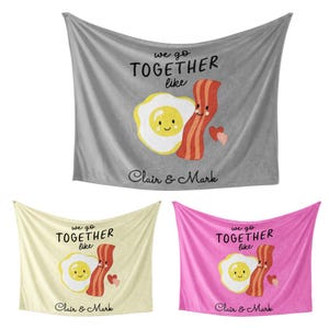 May include: Three fleece blankets featuring a cartoon illustration of a smiling egg and a smiling bacon strip. The text on the blankets reads "We go together like" and "Clair & Mark". The blankets are grey, yellow, and pink.