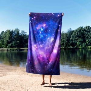 May include: A rectangular beach towel with a galaxy design in blue, purple, and white. The towel is held up against a backdrop of a lake and trees. The towel's design evokes a sense of the cosmos, perfect for outdoor use.