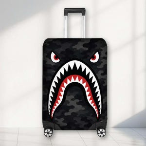 May include: A black and dark grey camouflage patterned suitcase featuring a graphic of a shark face with sharp white teeth, a red interior mouth, and two angry red and white eyes positioned above the mouth on the front of the luggage.