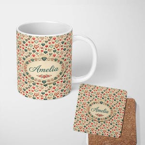 May include: A white ceramic mug and a square cork-backed coaster featuring a repeating pattern of small red, green, and beige hearts. Both items display a central oval label with a decorative border containing the name Amelia in green script.