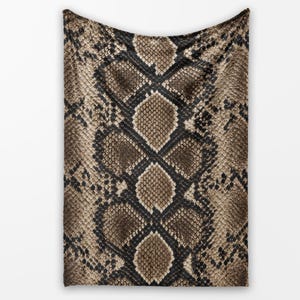 May include: A rectangular throw blanket featuring a repeating snakeskin pattern in shades of tan, brown, and black. The design displays a realistic scale texture with a central diamond-shaped motif running vertically across the fabric surface.