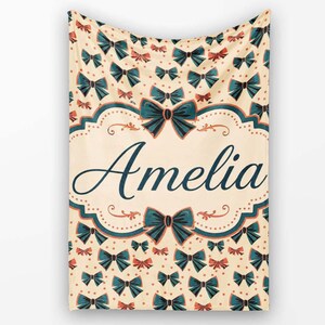 May include: A cream-coloured throw blanket featuring a repeating pattern of dark teal bows and small orange dots. The centre displays a decorative frame with the name Amelia written in a dark teal cursive script, flanked by two large bows.