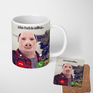 May include: White ceramic mug and coaster set featuring a pig-faced person in a plaid shirt with the text "John Pork is calling...". The mug has a curved handle and a scenic background.