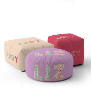 May include: Three colourful bean bag ottomans with personalised names. The first ottoman is beige with the name "ANDY" in pink and green letters. The second ottoman is purple with the name "LIZ" in green and pink letters. The third ottoman is red with the name "LUCY" in white letters.