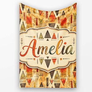 May include: A rectangular blanket featuring a cream-coloured central label with the name Amelia written in a large, cursive, multi-coloured font. The background is decorated with a repeating pattern of triangles and geometric shapes in orange, brown, and tan.