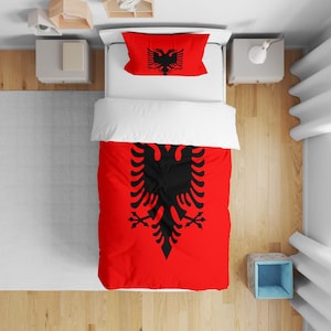 May include: A red and black bedding set with the Albanian flag design. The set includes a duvet cover and pillowcase, both showing the double-headed eagle emblem. The bedding is on a white bed, with a wooden floor visible.