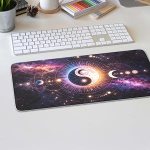 May include: A rectangular mouse mat with a cosmic design. The central feature is a Yin and Yang symbol, surrounded by a galaxy of purple, pink, and orange hues. A white keyboard is visible in the background, with a cup and stationery.