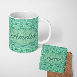 May include: A white ceramic mug and a square cork-backed coaster, both featuring a textured green and blue watercolour-style pattern. Each item displays the name Amelia in a dark green cursive script centred within a decorative scalloped frame.
