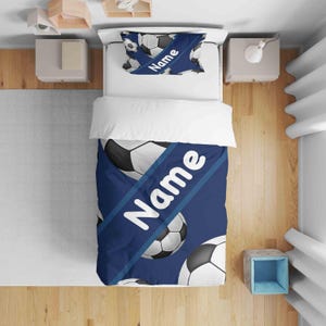 May include: A blue and white football-themed bedding set. The set includes a duvet cover and pillowcase, both featuring a repeating pattern of footballs and the word "Name" in white. The bedding is designed for a child's bed.