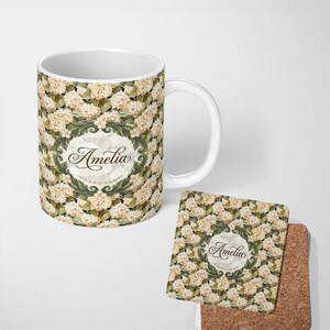 May include: A white ceramic mug and a square coaster featuring a repeating pattern of cream-coloured hydrangea flowers and green leaves. Both items display a central oval frame with an ornate green border containing the name Amelia in black script.