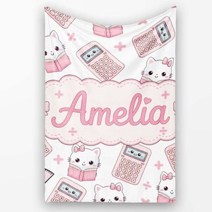 May include: A white blanket featuring a repeating pattern of pink calculators and white cats reading books. In the centre, a large pink scalloped label displays the name Amelia in a cursive font, surrounded by small pink plus and division signs.