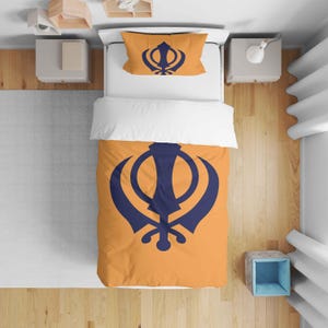 May include: Bedding set with an orange duvet cover and pillowcase, both displaying a navy blue Khanda symbol. The bedding is on a white bed, with a wooden floor visible. The set includes a duvet cover and a pillowcase.