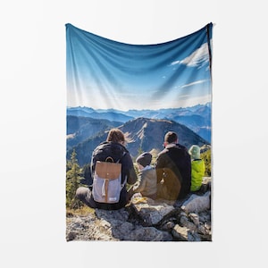 May include: A fleece blanket depicting a mountain scene with a family of four seated on a rocky outcrop. The blanket shows a blue sky and mountain range. The family members are wearing jackets and backpacks.