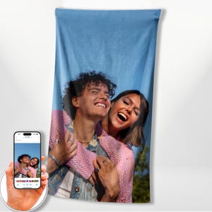 May include: A rectangular beach towel featuring a printed photograph of two people laughing together. One person wears a pink knit sweater over a denim jacket, while the other wears a pink shirt. The background is a clear blue sky.