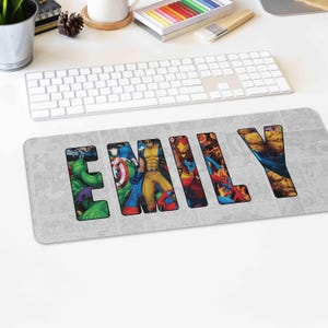 May include: A rectangular grey mouse mat featuring the name "EMILY" in large, bold letters. Each letter is filled with a different Marvel comic book character, including Hulk, Captain America, Wolverine, Iron Man, and The Thing.