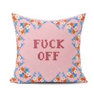 May include: Pink square cushion with cross-stitched text reading "FUCK OFF" in maroon. The cushion features a floral border in blue, orange, and white, offering a decorative and bold home decor piece.