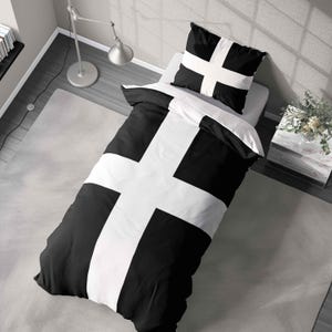 May include: A monochrome bedding set with a bold white cross pattern on a black duvet cover and pillowcase. The bed is neatly made, set against a neutral grey wall. The room has a minimalist design, with a rug and a side table.