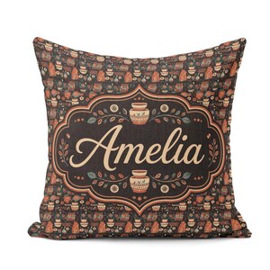 May include: A square throw pillow featuring a dark brown background with a repeating pattern of small pottery vessels. The centre displays a decorative frame containing the name Amelia in a light-coloured script font, flanked by two pottery icons.