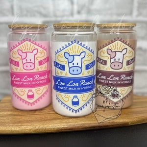 May include: Three glass tumblers with bamboo lids, each filled with a different colored liquid: pink, white, and tan. Each tumbler features a label with a cow graphic and the text "Lon Lon Ranch Finest Milk in Hyrule."