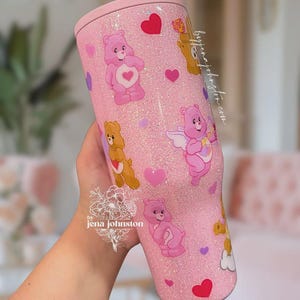 May include: A pink glitter tumbler featuring Care Bears characters and heart accents. The tumbler has a light pink lid and is decorated with various Care Bears in different poses. The design includes the text "by jena johnston".