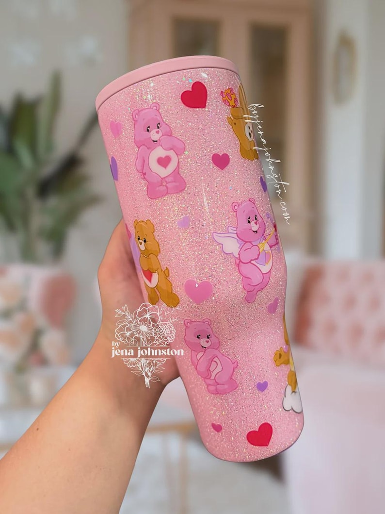 May include: A pink glitter tumbler featuring Care Bears characters and heart accents. The tumbler has a light pink lid and is decorated with various Care Bears in different poses. The design includes the text "by jena johnston".