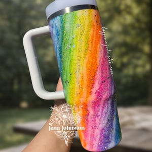 May include: A rainbow-colored glitter tumbler with a white handle and lid. The tumbler features a vibrant blend of colors, including pink, orange, yellow, green, blue, and purple. The handle is rectangular and the lid is white. The tumbler is cylindrical.