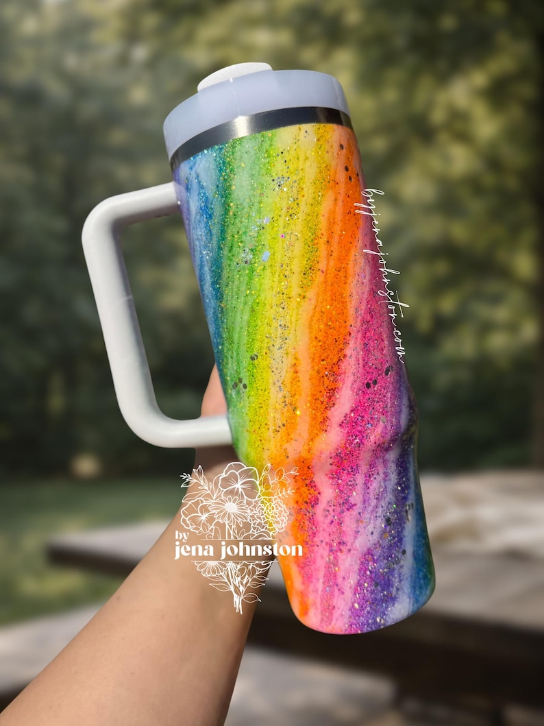 May include: A rainbow-colored glitter tumbler with a white handle and lid. The tumbler features a vibrant blend of colors, including pink, orange, yellow, green, blue, and purple. The handle is rectangular and the lid is white. The tumbler is cylindrical.