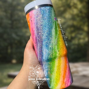 May include: A rainbow-colored tumbler with glitter accents. The tumbler features a gradient of colors, including pink, orange, yellow, green, blue, and purple. The text "by jena johnston" is visible on the tumbler.
