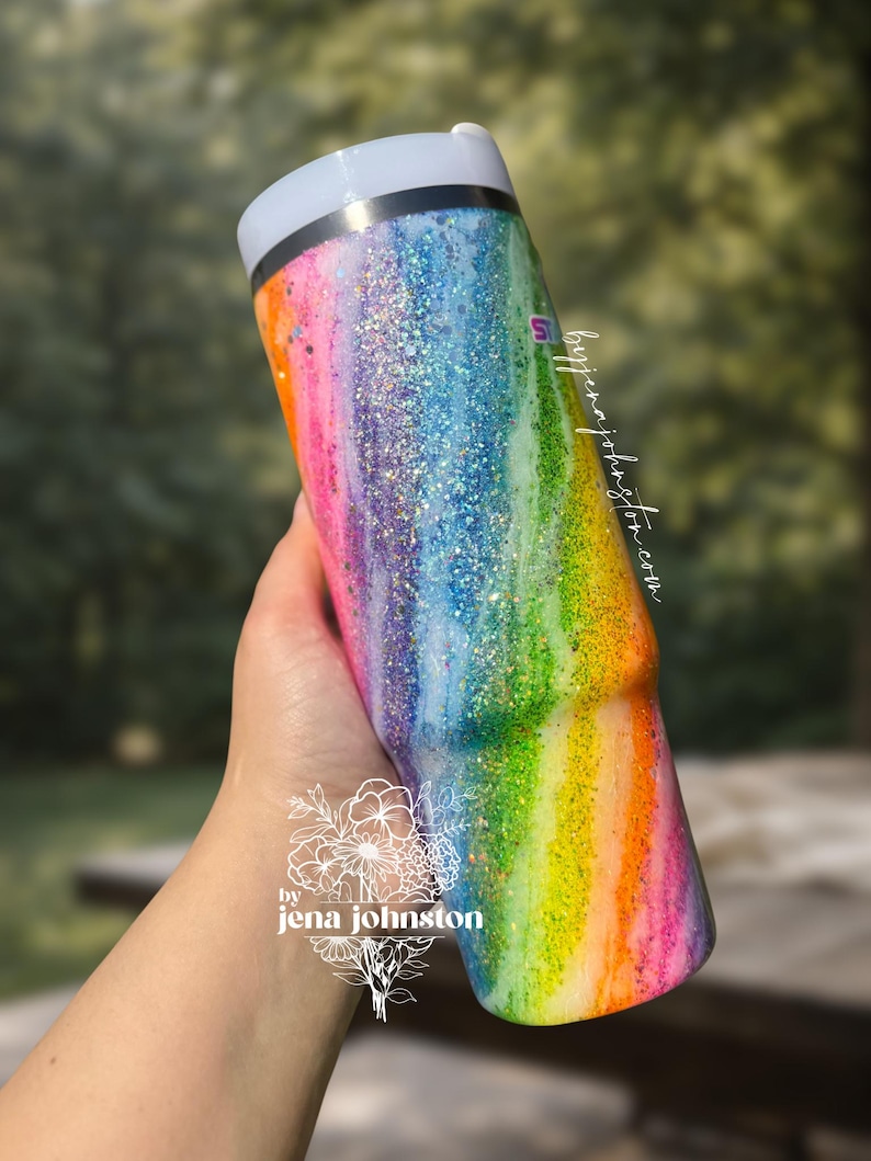May include: A rainbow-colored tumbler with glitter accents. The tumbler features a gradient of colors, including pink, orange, yellow, green, blue, and purple. The text "by jena johnston" is visible on the tumbler.