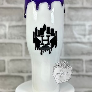 May include: White tumbler with a purple drip design at the top. The tumbler features a black silhouette of a city skyline with a star and the letter "H". The bottom of the tumbler has the text "by jena johnston".