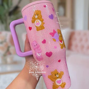 May include: Pink glitter tumbler with a light purple handle and lid. The tumbler is decorated with Care Bear characters and hearts. The tumbler has the text "by jena johnston" on it.