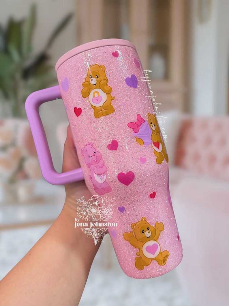 May include: Pink glitter tumbler with a light purple handle and lid. The tumbler is decorated with Care Bear characters and hearts. The tumbler has the text "by jena johnston" on it.