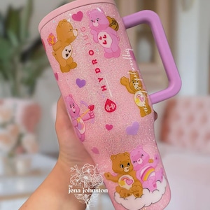 May include: Pink glitter tumbler with Care Bear graphics, a light purple handle, and a matching lid. The tumbler features various Care Bears, hearts, and the text "HYDRO JUG". The tumbler is held by a person.