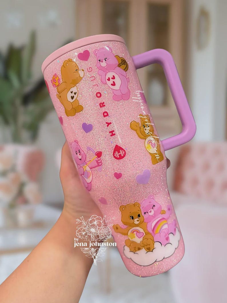 May include: Pink glitter tumbler with Care Bear graphics, a light purple handle, and a matching lid. The tumbler features various Care Bears, hearts, and the text "HYDRO JUG". The tumbler is held by a person.