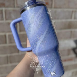 May include: A lavender glitter tumbler with a handle and a clear lid. The tumbler has a marbled white design and a small white sticker with a winged dog. The text "by jena johnston" is visible on the side.