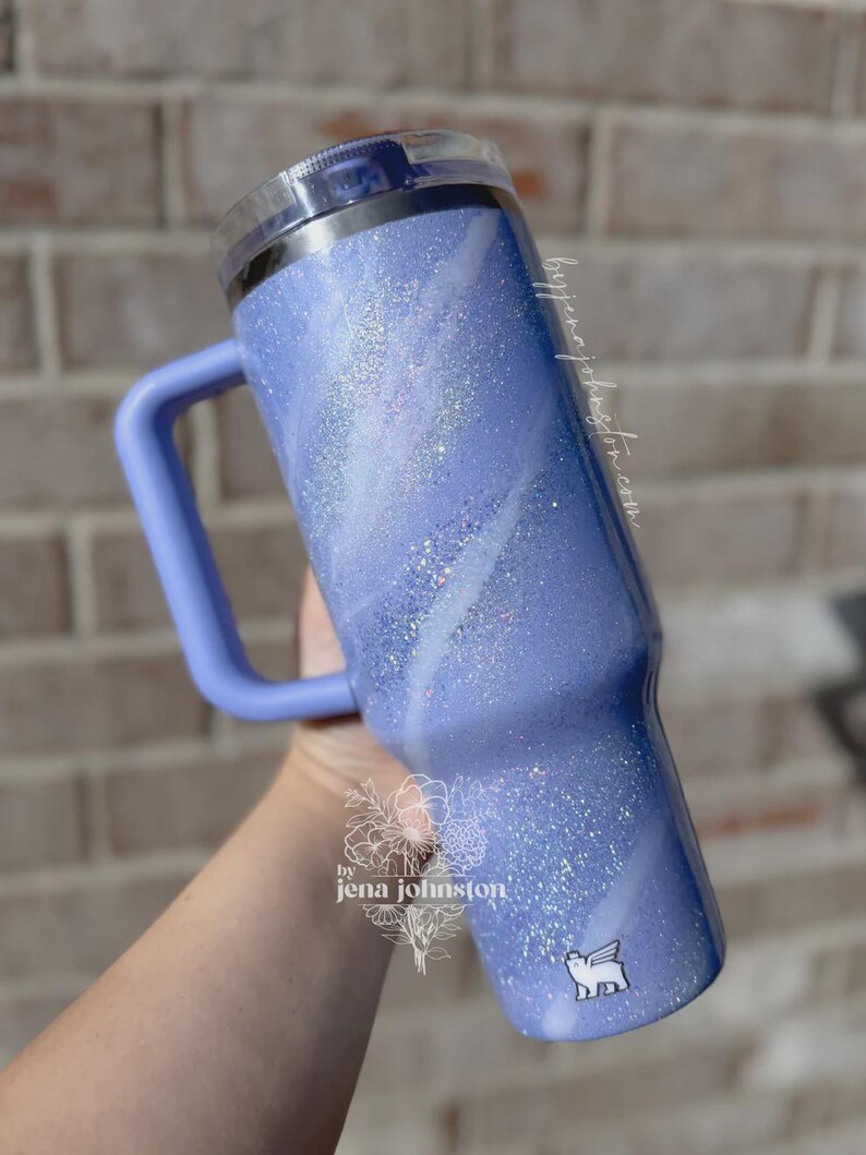May include: A lavender glitter tumbler with a handle and a clear lid. The tumbler has a marbled white design and a small white sticker with a winged dog. The text "by jena johnston" is visible on the side.