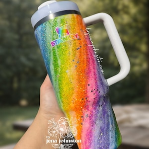 May include: A rainbow-colored Stanley tumbler with a white handle and lid. The tumbler has a glittery finish and features the Stanley logo. The text "by jena johnston" is also visible.