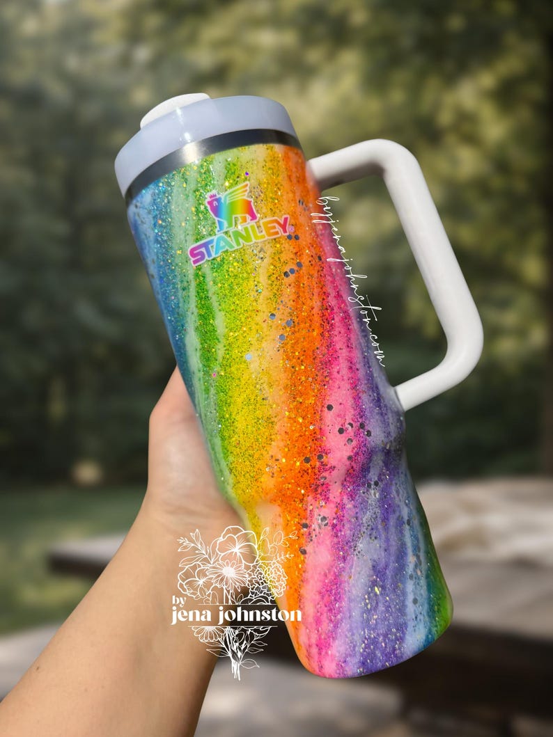 May include: A rainbow-colored Stanley tumbler with a white handle and lid. The tumbler has a glittery finish and features the Stanley logo. The text "by jena johnston" is also visible.