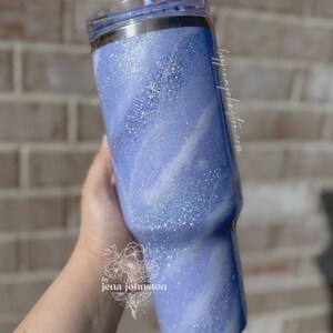 May include: A blue glitter tumbler with a swirling white design. The tumbler has a clear lid and is held by a hand. The text "by jenajohnston.com" is visible on the side. The tumbler is a stylish drinkware item.